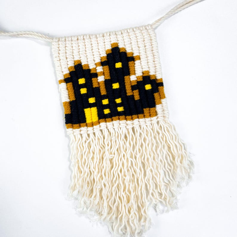 May include: A white macrame wall hanging with a black and yellow cityscape design. The hanging has a fringe border.