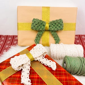 K&ouml;nnte beinhalten: Zwei Geschenkboxen, die in braunes Papier mit goldenem Band und gr&uuml;n-wei&szlig;en H&auml;kelb&ouml;gen verpackt sind. Eine Box liegt auf der anderen, und im Vordergrund befindet sich eine Spule mit wei&szlig;em Garn und eine Spule mit gr&uuml;nem Garn.