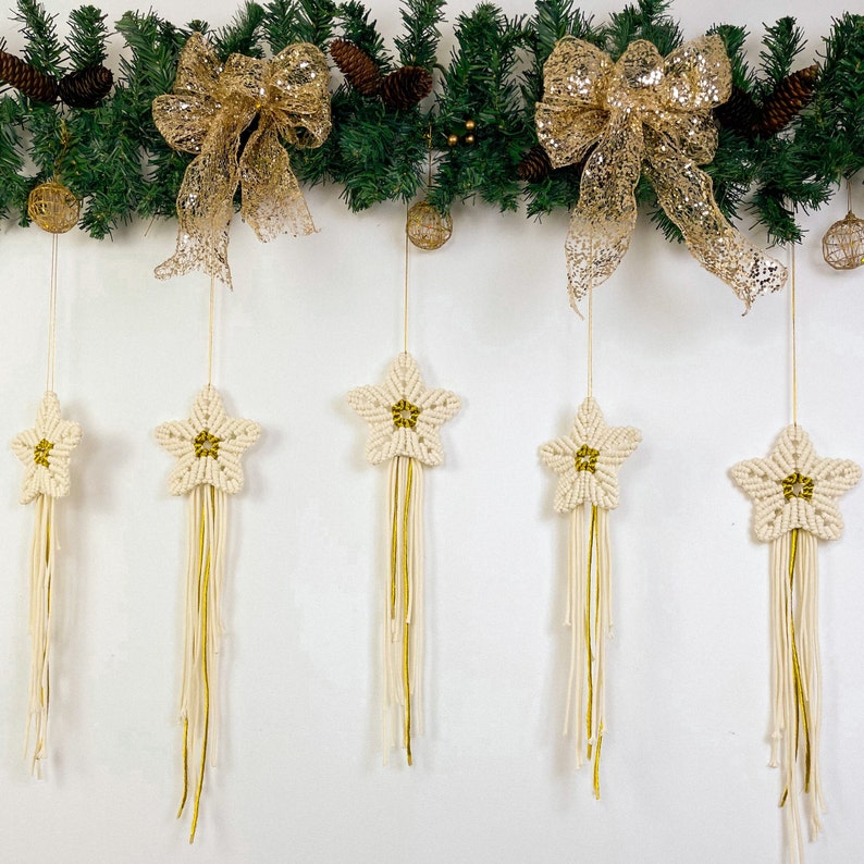 May include: Five white macrame star ornaments with gold accents hanging from a garland of green pine branches with gold bows.