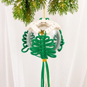 Op de afbeelding: Een groene en witte gehaakte kerstboomversiering met een gouden ster bovenop. De versiering hangt aan een tak van een kerstboom.
