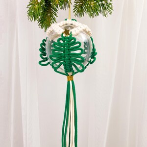 Op de afbeelding: Een groene en witte macram&eacute; kerstversiering in de vorm van een boom die aan een dennentak hangt. De versiering is gemaakt van een heldere glazen bal en een lange kwast.