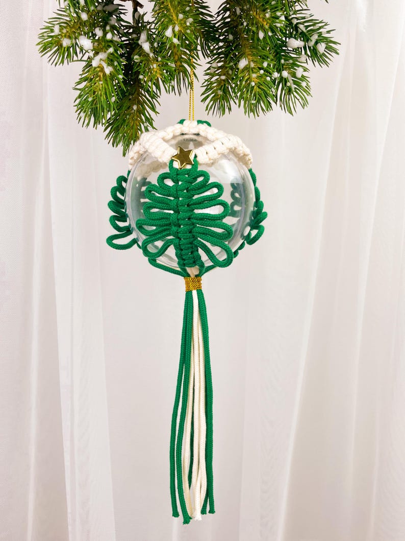 Op de afbeelding: Een groene en witte macram&eacute; kerstversiering in de vorm van een boom die aan een dennentak hangt. De versiering is gemaakt van een heldere glazen bal en een lange kwast.