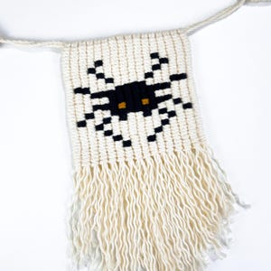 May include: A white woven wall hanging with a black spider design and yellow eyes. The hanging has a fringe border.