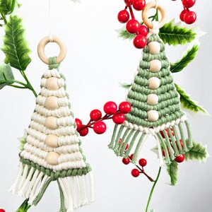 May include: Two handmade macrame Christmas tree ornaments with wooden beads. The ornaments are green and white with fringe. The ornaments are hanging from a branch with red berries.