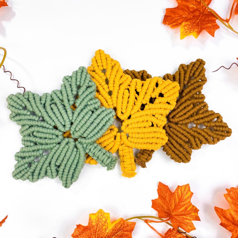 Op de afbeelding: Drie macram&eacute; wandhangers in de vorm van bladeren in groen, geel en bruin. De bladeren zijn aan elkaar gebonden en hangen tegen een witte achtergrond.