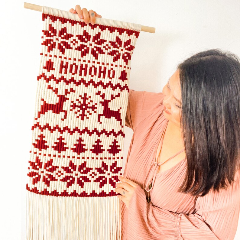 May include: A white and red knitted wall hanging with a Christmas theme. The hanging features a snowflake, reindeer, and Christmas trees. The text "HOHOHO" is woven into the design.