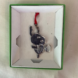 May include: A silver ornament of a cartoon dog holding a ball, hanging from a red ribbon in a green box.