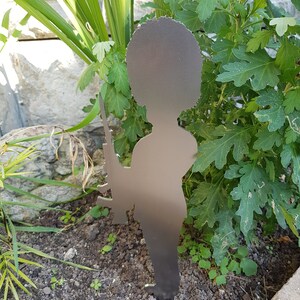 May include: A metal garden ornament depicting a soldier with a rifle. The dark grey silhouette is positioned in a garden bed, surrounded by green foliage and a stone wall. The stake is designed to be inserted into the soil.