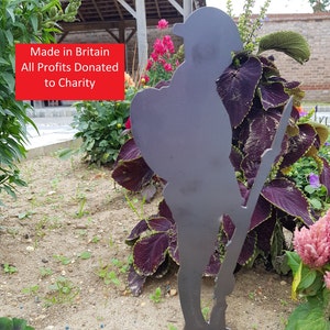 May include: A metal silhouette of a soldier standing in a garden with purple and green plants. The silhouette is made of dark gray metal and is holding a rifle. The text "Made in Britain All Profits Donated to Charity" is visible in the image.
