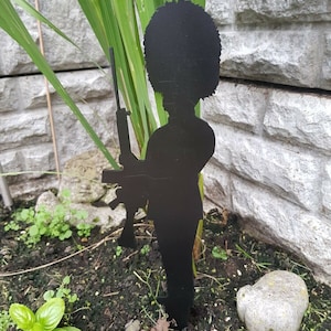 May include: Black metal silhouette of a British guard standing at attention, holding a rifle. The silhouette is placed in a garden setting with green plants and brown soil.