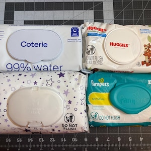 May include: Four packages of baby wipes are displayed. Brands include Coterie, Huggies, Pampers, and a brand with a star pattern. The Coterie wipes are labeled "99% water". The Huggies package features a cartoon tiger.