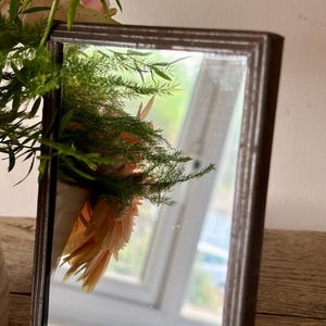 May include: A rectangular mirror with a dark brown frame reflecting a bouquet of peach-coloured flowers and green foliage. The mirror surface shows a clear reflection of the floral arrangement and a window in the background, capturing light.