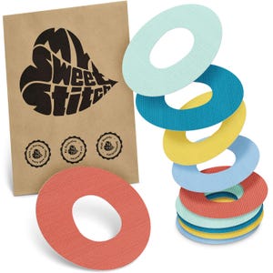 Könnte beinhalten: Mehrfarbige, strukturierte, ovale Klebepflaster in Blau-, Gelb- und Rottönen. Im Hintergrund ist eine braune Papiertüte mit dem Text "My Sweet Stitch" und drei kleinen runden Logos zu sehen.