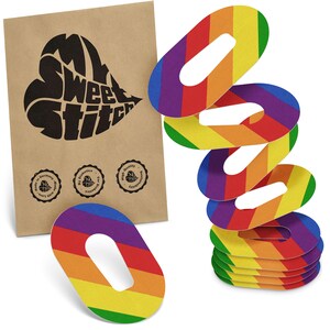 May include: A collection of rainbow-striped oval stickers with a central opening, displayed beside a brown paper packet. The stickers showcase a vibrant array of colours. The packet features the text "My Sweet Stitch" in a heart shape and small circular logos.