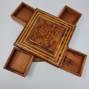 May include: A square wooden box with a central lid featuring a dark brown burl wood grain pattern. The lid is framed by thin, light-colored inlay lines. Four open, square-shaped drawers extend outward from beneath the central lid.