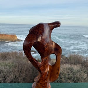 May include: A polished, reddish-brown wooden sculpture featuring a complex, organic shape with two distinct open voids. The piece is mounted on a circular wooden base and displays a rich, swirling grain pattern throughout its smooth, curved surface.