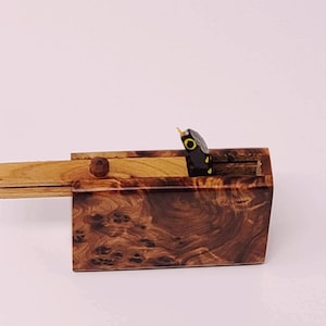 May include: A wooden box with a hinged lid, featuring a small black and yellow snake figurine peeking out from the top. The box is made of a dark wood with a unique grain pattern.
