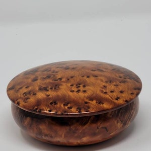 May include: A round wooden box with a lid. The box is made of a light brown wood with a dark brown swirl pattern. The lid is closed.