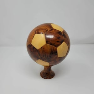 May include: A wooden soccer ball ornament with a brown and yellow wood grain pattern. The ball is on a small wooden stand.