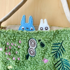 May include: A green textured tote bag with embroidered floral and leaf designs. Two cartoon rabbit-like characters peek over the top, one blue and one white. The bag has a beige strap and a light brown interior.