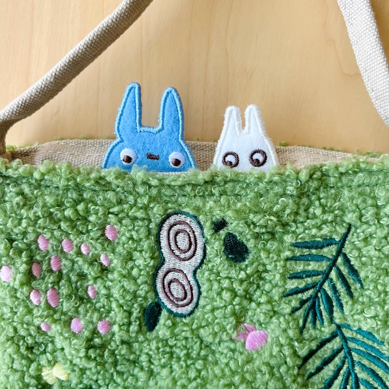 May include: A green textured tote bag with embroidered floral and leaf designs. Two cartoon rabbit-like characters peek over the top, one blue and one white. The bag has a beige strap and a light brown interior.