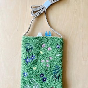 May include: A green textured crossbody bag with embroidered floral and leaf designs. The bag features a long, beige strap and a small, light blue accent at the top. The bag is decorated with blue, pink, and white floral embroidery.
