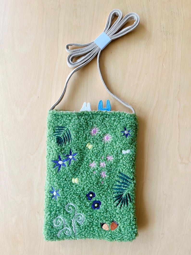 May include: A green textured crossbody bag with embroidered floral and leaf designs. The bag features a long, beige strap and a small, light blue accent at the top. The bag is decorated with blue, pink, and white floral embroidery.