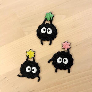May include: Three black embroidered patches depicting cartoon creatures with large white eyes and small legs. Each creature holds a colourful star above its head. The stars are green, pink, and yellow.