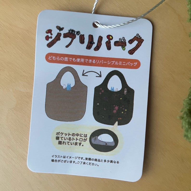 May include: A tag for a reversible mini bag featuring illustrations. One side is brown with a Totoro graphic, the other is black with floral and Totoro designs. Japanese text is visible, including "Ghibli Park" and descriptions.