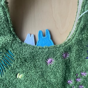 May include: Green plush tote bag with embroidered floral and leaf accents. Features two felt character appliqu&eacute;s in blue and white. The bag has a soft, textured appearance and a neutral-colored interior.