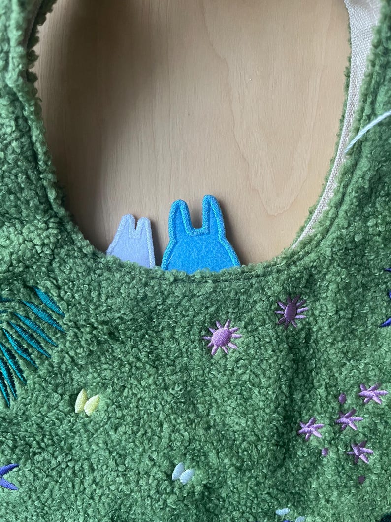 May include: Green plush tote bag with embroidered floral and leaf accents. Features two felt character appliqu&eacute;s in blue and white. The bag has a soft, textured appearance and a neutral-colored interior.