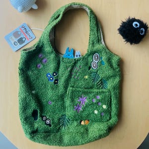 May include: Green tote bag with embroidered floral and leaf designs. The bag has a pocket and a plush texture. A tag with Japanese text is attached. A small blue bunny and a black plush character are also visible.