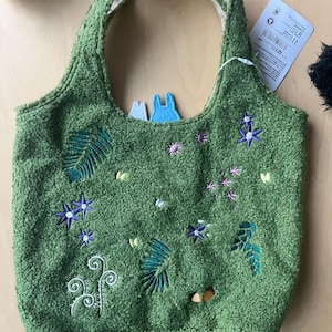 May include: Green tote bag with embroidered floral and leaf designs. The bag has a textured, plush appearance and a light-colored interior. Two small felt figures are visible inside the bag.