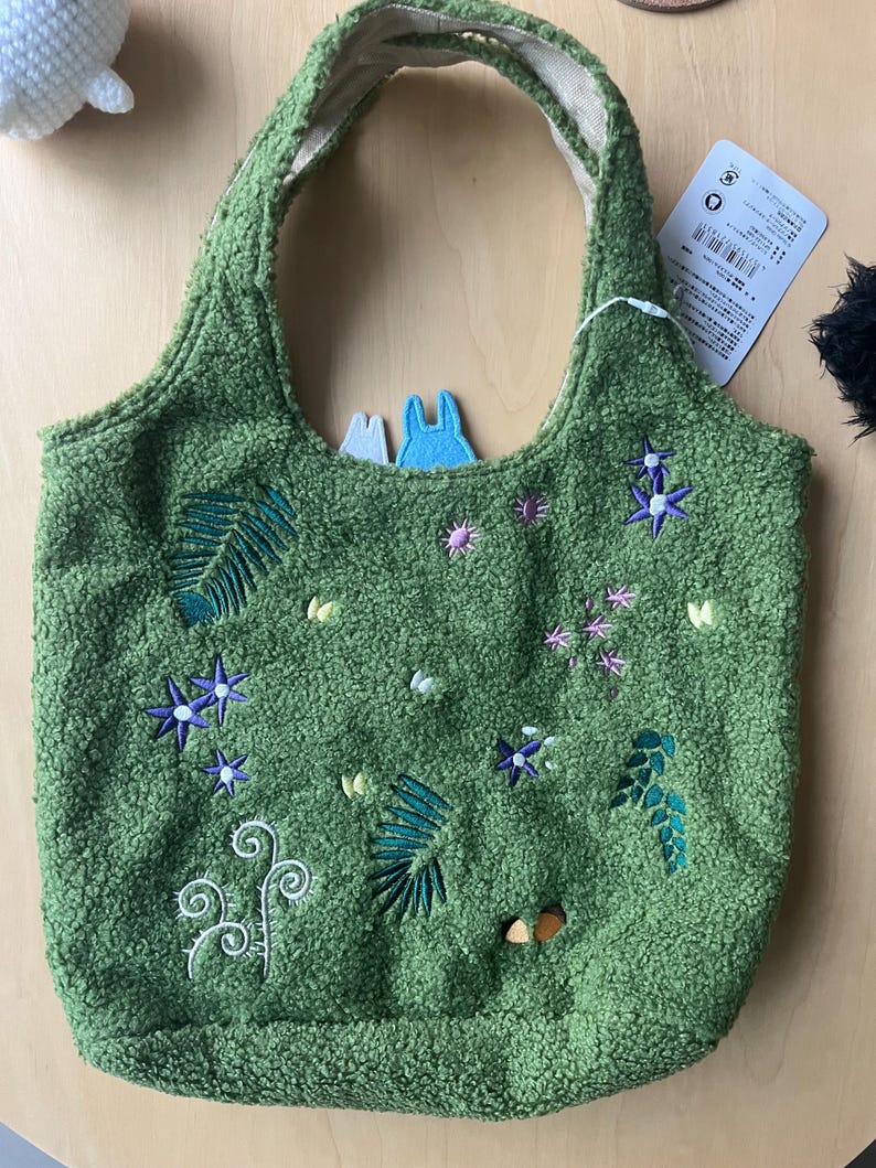 May include: Green tote bag with embroidered floral and leaf designs. The bag has a textured, plush appearance and a light-colored interior. Two small felt figures are visible inside the bag.