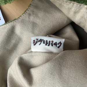 May include: Close-up of a green and tan tote bag. The bag has a soft, textured green trim and a tan canvas body. A small white tag with Japanese text is sewn onto the bag. The bag is partially open, revealing the interior.