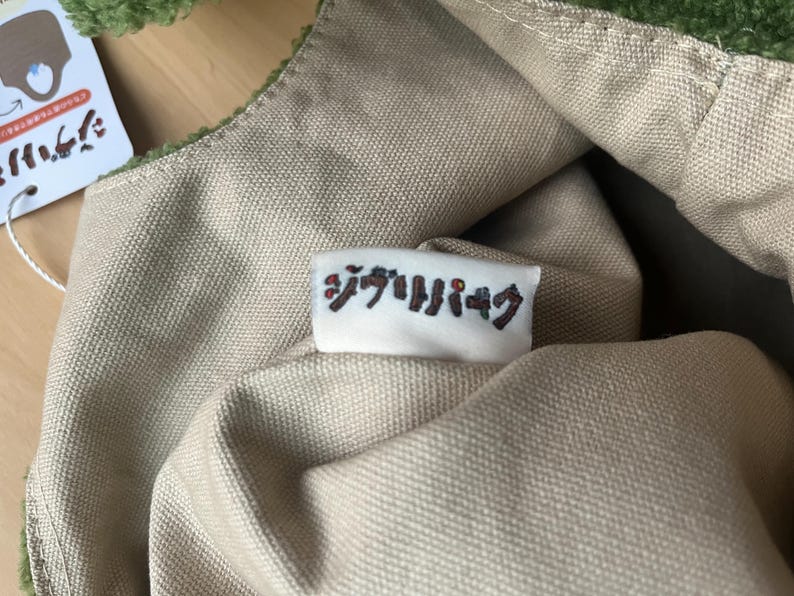 May include: Close-up of a green and tan tote bag. The bag has a soft, textured green trim and a tan canvas body. A small white tag with Japanese text is sewn onto the bag. The bag is partially open, revealing the interior.