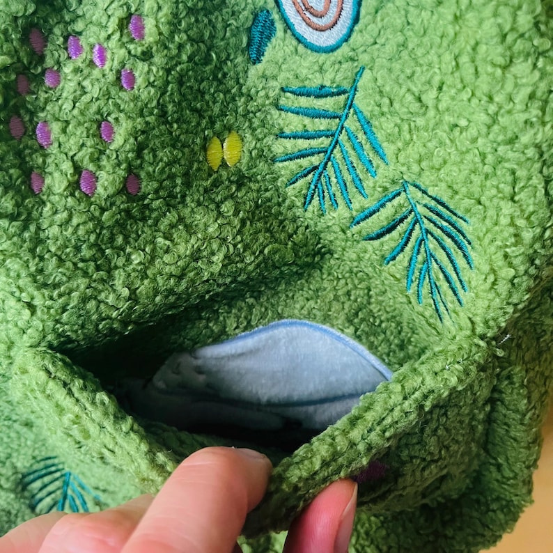 May include: Close-up of a green, textured fabric with embroidered nature-themed designs. Features a light blue pocket and decorative elements in teal, yellow, and purple. The fabric has a soft, plush appearance.