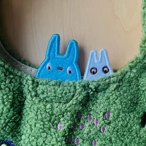 May include: A green, textured fabric bag with two blue bunny-shaped patches peeking out of a pocket. The patches have black eyes and embroidered details. The bag has a light gray lining and purple embroidered accents.