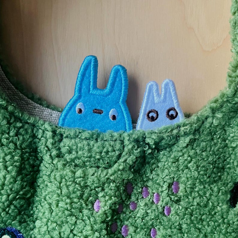 May include: A green, textured fabric bag with two blue bunny-shaped patches peeking out of a pocket. The patches have black eyes and embroidered details. The bag has a light gray lining and purple embroidered accents.