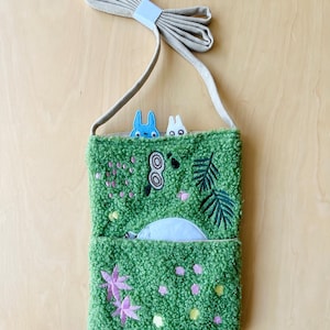 May include: A green textured crossbody bag with embroidered floral and leaf designs. The bag features a light blue zipper pocket and a beige strap. Two cartoon characters peek over the top. The bag is approximately 8 inches wide and 10 inches tall.