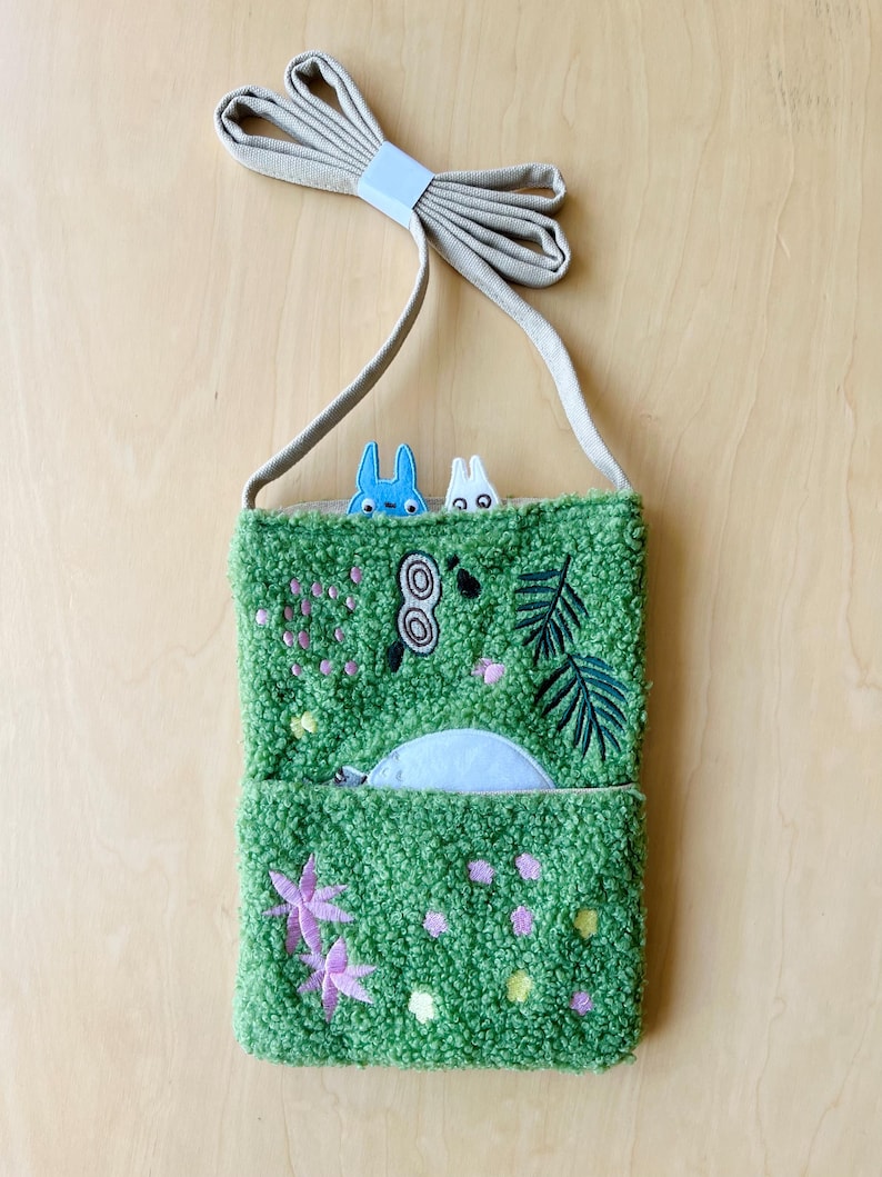 May include: A green textured crossbody bag with embroidered floral and leaf designs. The bag features a light blue zipper pocket and a beige strap. Two cartoon characters peek over the top. The bag is approximately 8 inches wide and 10 inches tall.