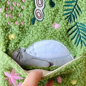 May include: Close-up of a green, textured fabric with embroidered floral and leaf designs. A gray plush animal with a white shell is nestled in a pocket. The pocket is lined with a beige fabric.