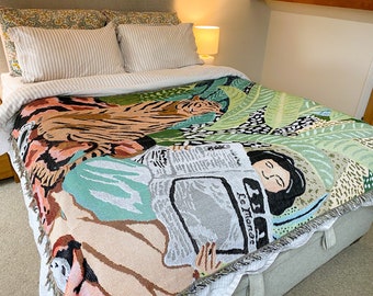 Puede incluir: Una manta de tiro de tapiz tejido presenta un diseño colorido de un tigre, una mujer leyendo un periódico y plantas tropicales. La manta tiene un borde con flecos.