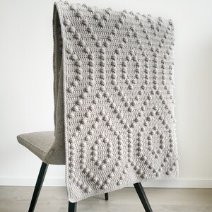 May include: A grey crocheted throw blanket with a textured pattern, draped over a black chair with a grey seat.