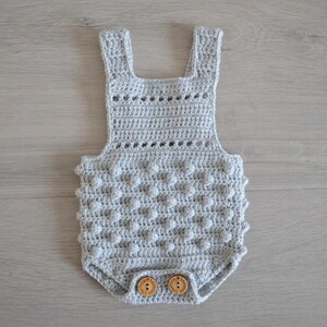 May include: A gray crocheted baby onesie with a textured, bobble stitch pattern. The onesie has a crocheted strap design and wooden buttons.