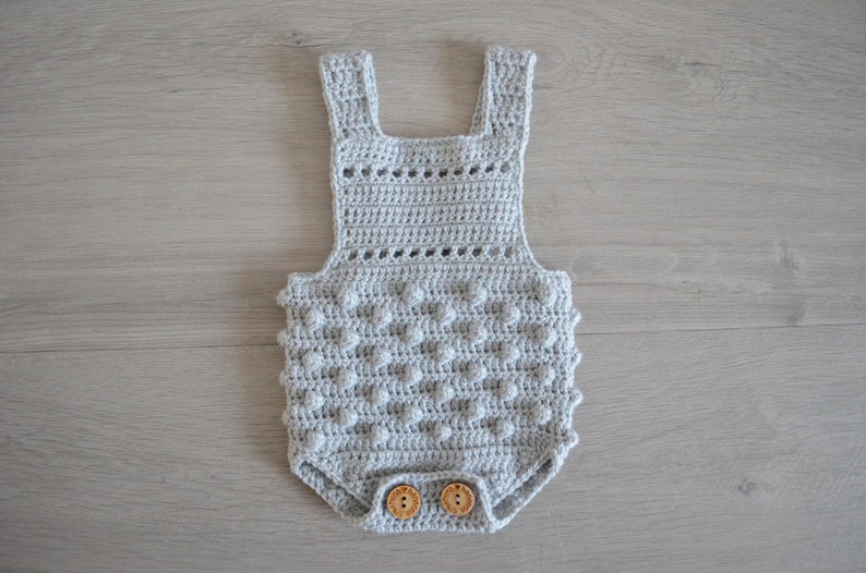 May include: A gray crocheted baby onesie with a textured, bobble stitch pattern. The onesie has a crocheted strap design and wooden buttons.