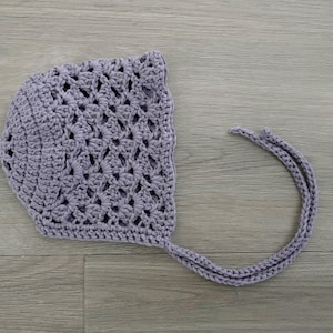 Peut inclure: Un bonnet de bébé en crochet de couleur mauve clair avec une longue attache. Le bonnet a un motif ajouré délicat.