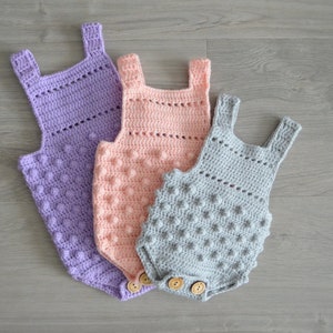 May include: Three crocheted baby onesies in different sizes. The onesies are made with a textured stitch and have a button closure. The onesies are in shades of purple, pink, and gray.