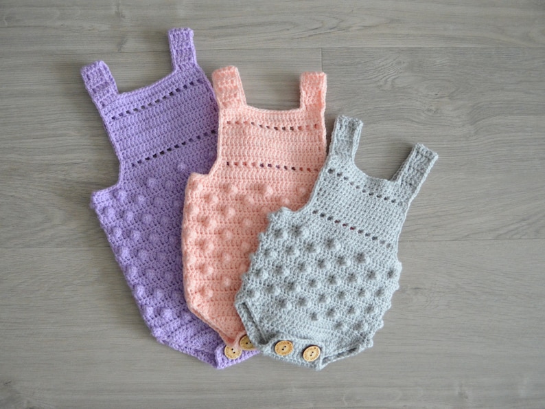 May include: Three crocheted baby onesies in different sizes. The onesies are made with a textured stitch and have a button closure. The onesies are in shades of purple, pink, and gray.