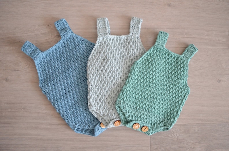 May include: Three crocheted baby onesies in different colors: light blue, gray, and green. Each onesie has a button closure and a textured pattern.